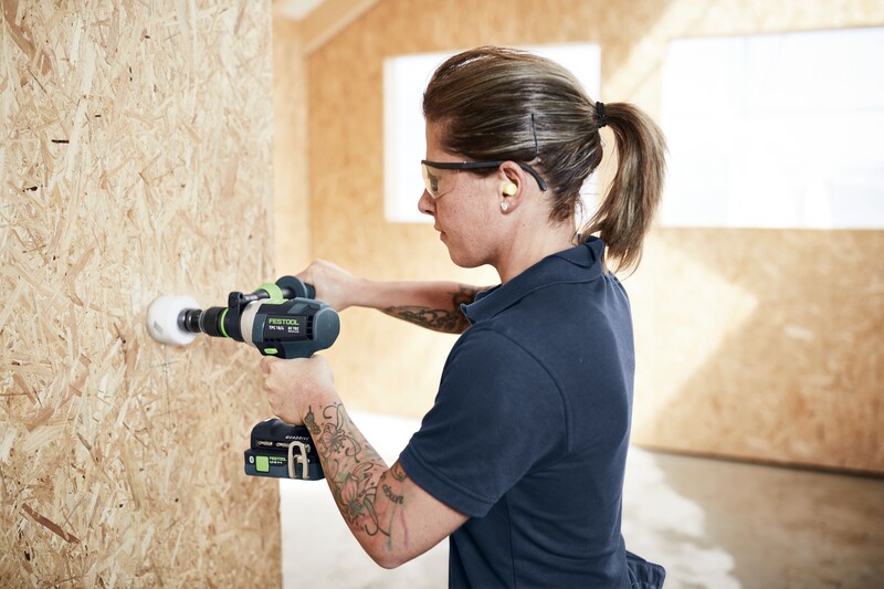 A person wearing safety glasses and earplugs uses a power drill with a hole saw attachment to drill into a wooden wall inside a brightly lit workshop.