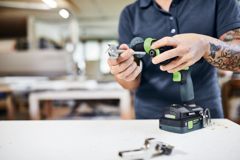 A person with tattooed arms assembles or adjusts a green and black cordless power drill on a white workbench in a workshop, with tools and a blurred background visible.