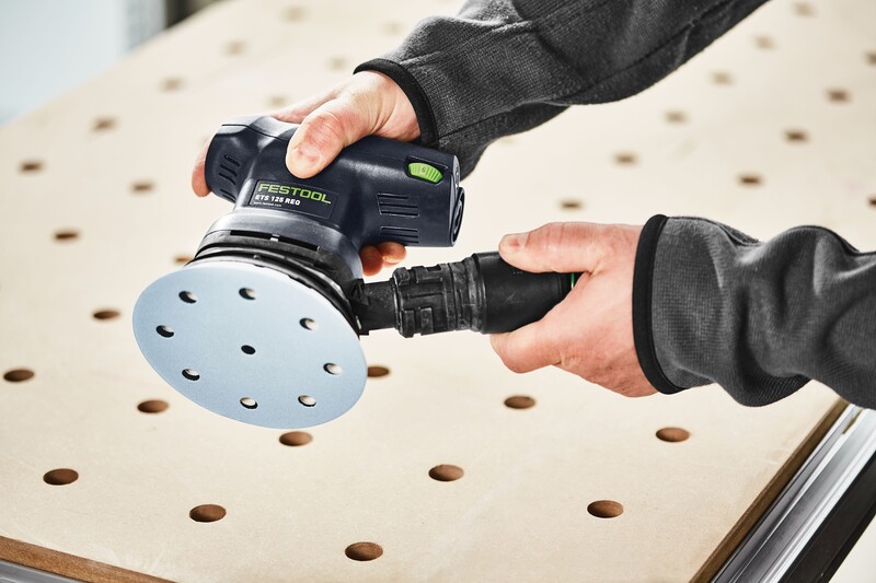A person holds a Festool orbital sander above a perforated workbench, preparing to sand a flat wooden surface.