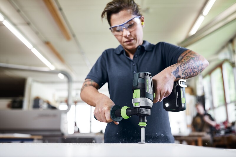 A person wearing safety glasses uses a power drill on a workbench in a workshop. They have tattoos on their arms and are focused on the task. Bright natural light comes through the large windows in the background.