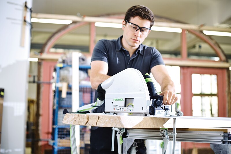 A man wearing safety glasses and gloves uses a Festool circular saw to cut a piece of wood on a workbench in a bright, industrial workshop.