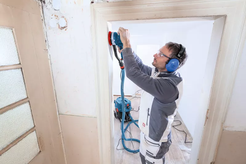 A man wearing safety glasses and ear protection uses a Bosch 0601257170 GET 75-150 Professional Turbo Sander 240v to sand a door frame in a room under renovation, with tools and extension cords visible on the floor.