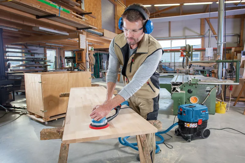 Wearing safety glasses and ear protection, a man uses the Bosch 0601257170 GET 75-150 Professional Turbo Sander 240v on a wooden board in a workshop filled with woodworking tools and machines.