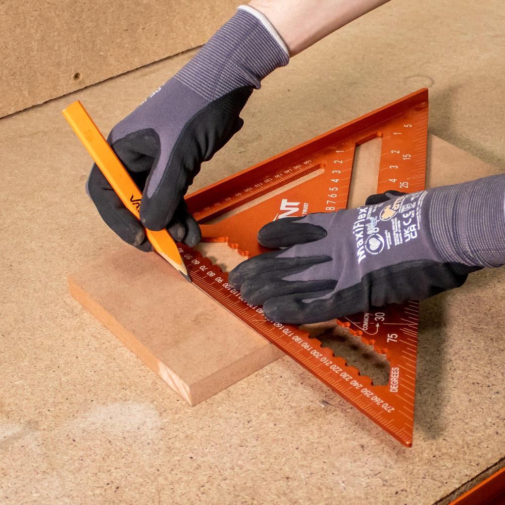 A person wearing black and gray gloves marks a piece of wood with an orange pencil, using an orange triangular ruler for measurement on a workbench.