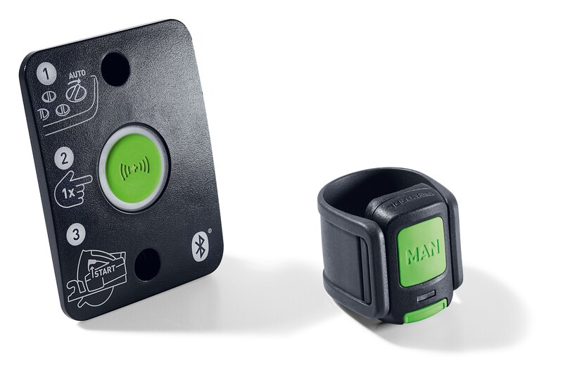 A black control panel with a large green button and illustrations, next to a black and green wearable ring with MAN written on its button, likely used for remote operation or automation.