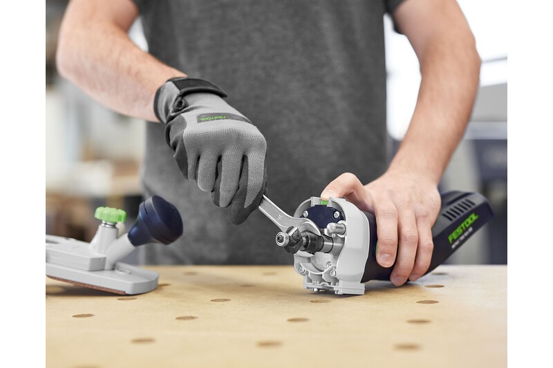 Wearing gray gloves and a gray shirt, a person uses a wrench to adjust a blue and silver Festool 578120 Cordless Joining Machine DOMINO DFC 500 E-Basic on a wooden workbench, with another tool attachment resting nearby.