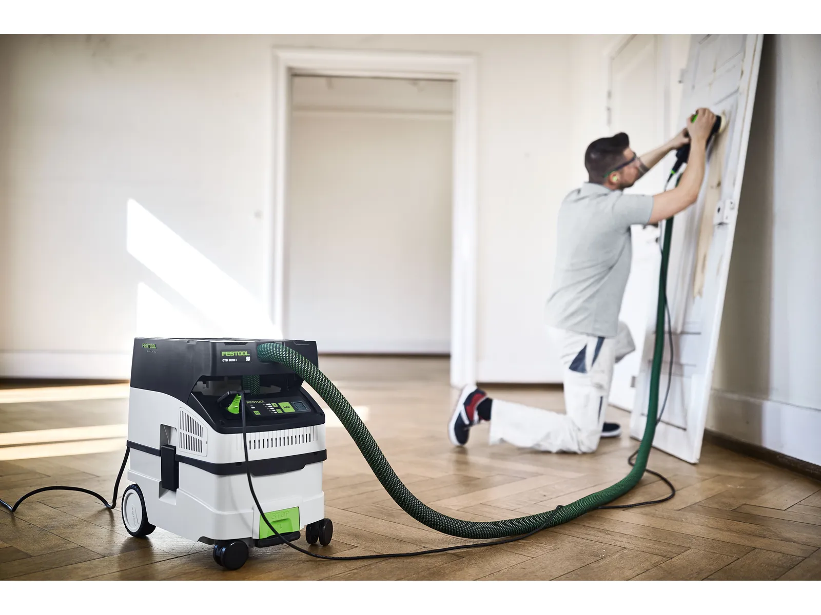 A person kneels while sanding a white door attached by a green hose to a Festool 578321 CTL MIDI I 230V Mobile Dust Extractor CLEANTEC on a wooden floor in a bright, empty room.