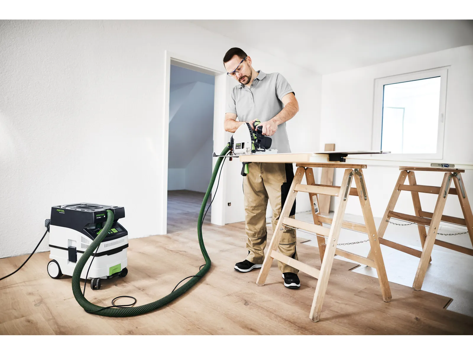 A man uses a circular saw connected to a Festool 578321 CTL MIDI I 230V Mobile Dust Extractor CLEANTEC on wood supported by two sawhorses in a bright, unfinished room with wooden flooring.