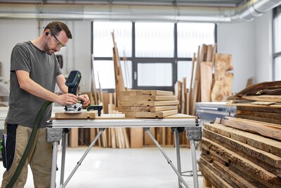 A man with glasses and ear protection uses the Festool 578120 Cordless Joining Machine DOMINO DFC 500 E-Basic to work on wood in a bright, organized workshop with stacks of wood and large windows in the background.