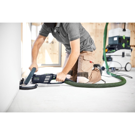 A person kneels on the floor using the RG130 ECI-PLUS 230V with a green hose to sand near a wall, vacuum device in background.