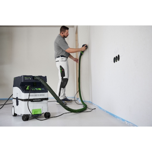 A man sands a white wall in a minimally furnished room using a power sander attached to a Festool dust extractor with a green hose.