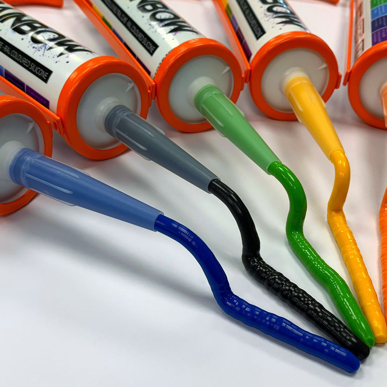 Five tubes of colored sealant lie on a white surface with blue, gray, black, green, and yellow sealant squeezed out in curved lines.
