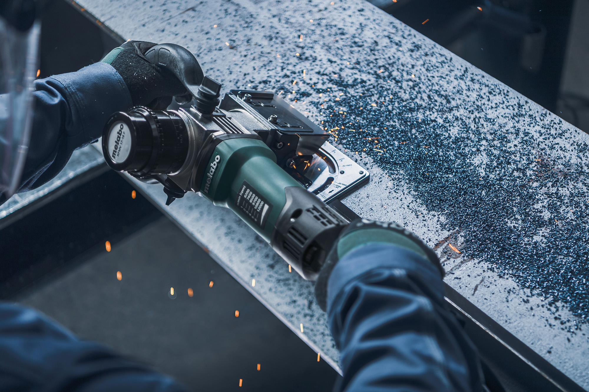 A person in protective gloves uses a handheld power grinder on a metal beam, sparks and shavings scattering across the surface.