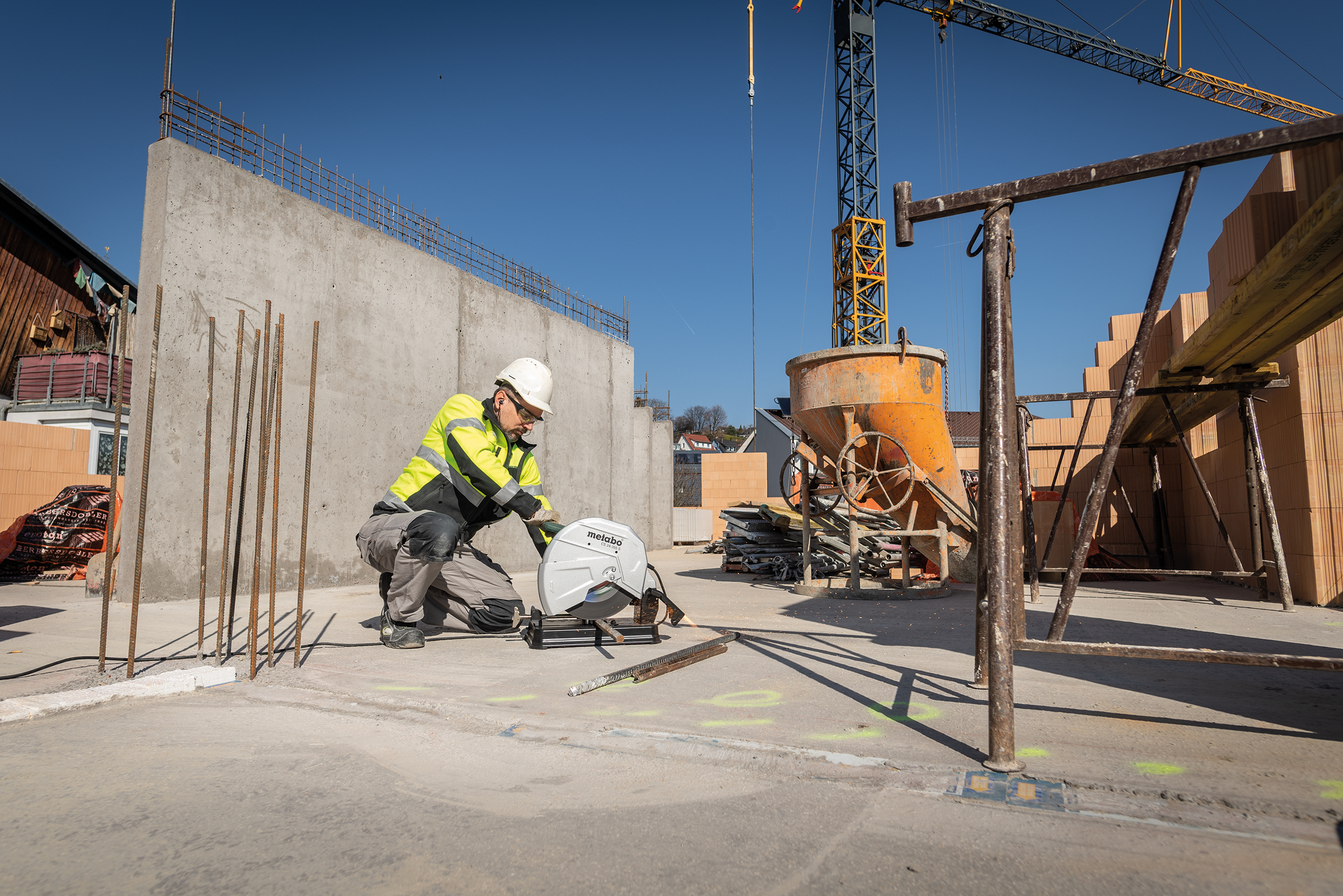 A construction worker in a high-visibility jacket and hard hat saws metal rods at a building site with concrete, bricks, and a crane.