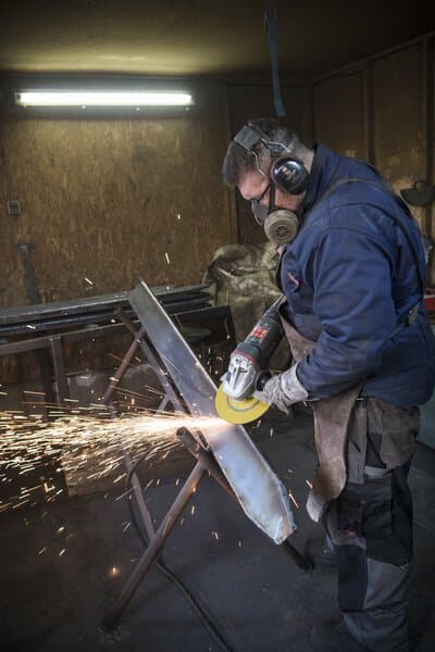 Wearing safety gear, a person grinds a metal sheet in a workshop with an angle grinder, bright sparks flying around.