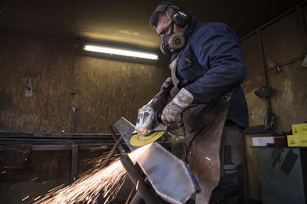 Wearing protective gear, a person uses a grinding disc on metal, producing bright sparks in a dimly lit workshop.