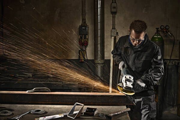 Wearing safety gear, a person grinds a metal beam with an angle grinder, creating sparks in a workshop filled with tools.