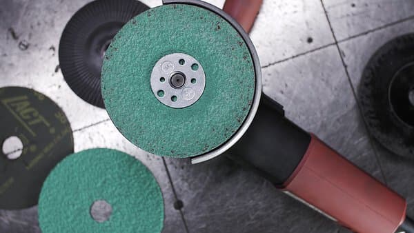 A close-up of an angle grinder with a 316492 FS966 115x22 60 grit fibre Ceramic Multibond disc on a metallic surface, extra discs nearby.