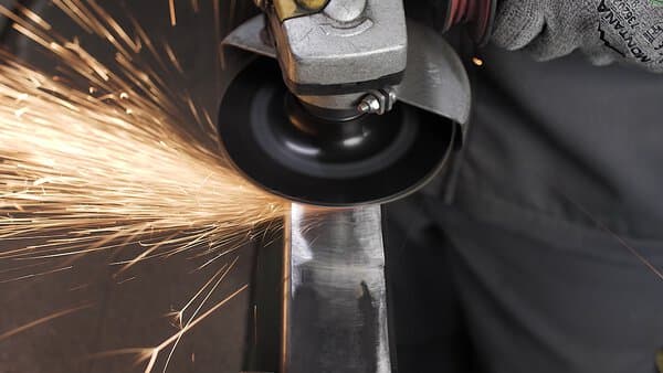 A person uses a 60 grit fibre disc on an angle grinder to smooth metal, bright sparks flying as they work. They wear gloves.