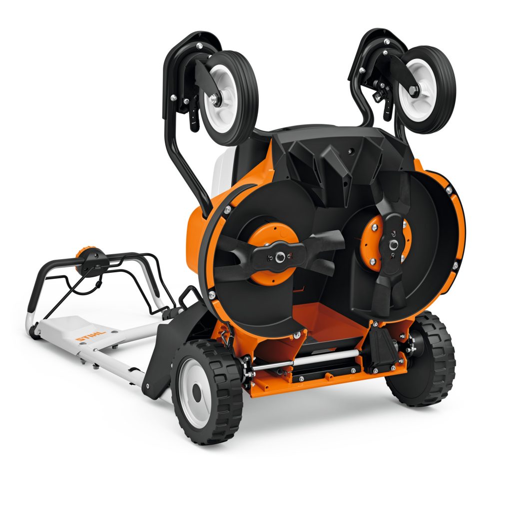 A Stihl RMA 765.0 V (AR) Lawn Mower tipped sideways displays two rotary blades, a black and orange body, and large wheels facing upwards.