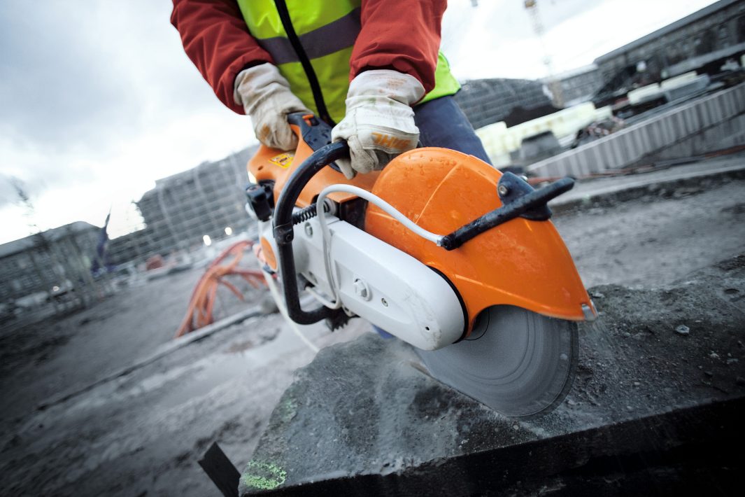 A construction worker in gloves and a high-visibility jacket uses a cut-off saw on concrete at a site with buildings and equipment.