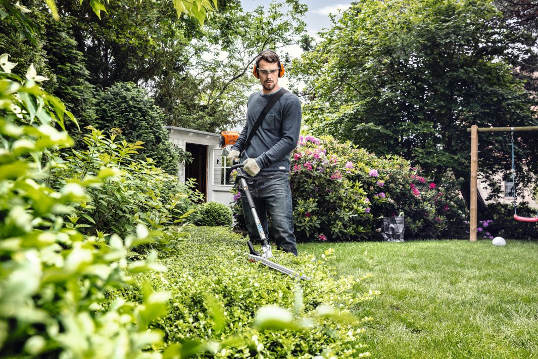 A man in gloves and casual clothes trims bushes in a green garden with a hedge trimmer; shrubs, swing set, and shed behind.