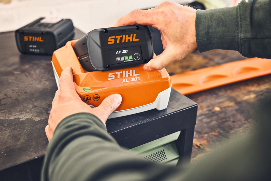 A person places a STIHL AP 20 battery on an orange and white STIHL AL 301 charger, with another battery and tools in the background.