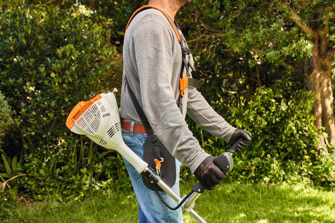 Wearing gloves and a harness, a person operates the Stihl FS 56 C-E Brushcutter in a sunny garden with bushes and trees.
