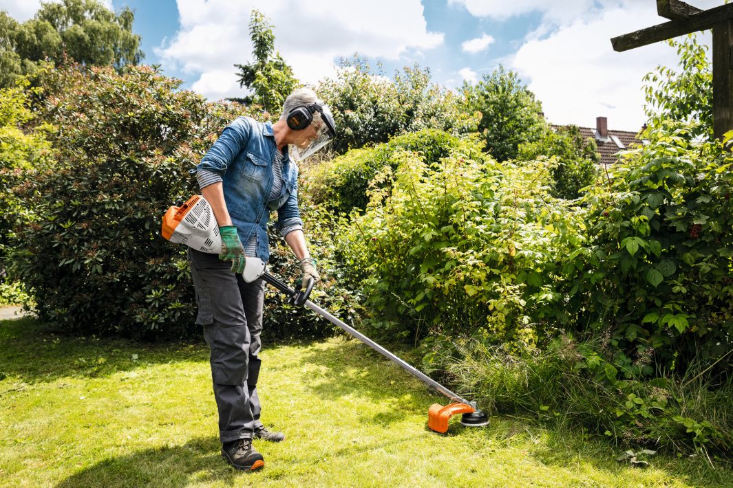 Wearing protective gear, a person trims grass along a lush garden edge with a strimmer as trees and bushes surround the sunny garden.