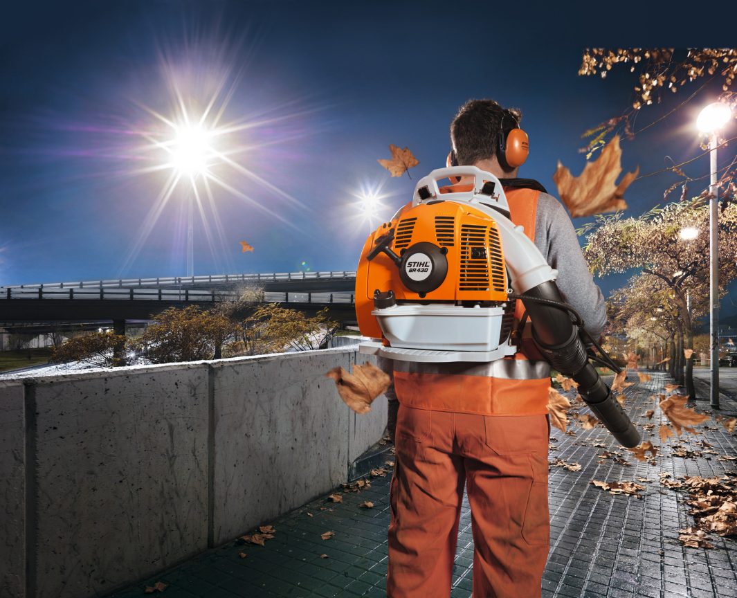 Wearing safety gear, a person uses a Stihl BR 430 Backpack Blower at night to clear autumn leaves from a lit walkway with trees and streetlights.