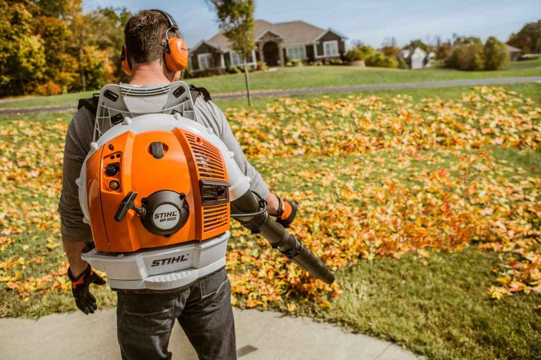 Wearing ear protection, a person uses the Stihl BR 600 Backpack Blower to clear fallen leaves from a garden near a house and trees.