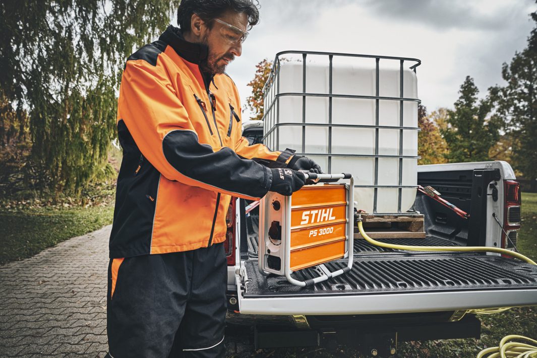 Wearing orange and black protective gear, a person handles a Stihl PS 3000.0 on the tailgate of a pickup with a large white container in the back.