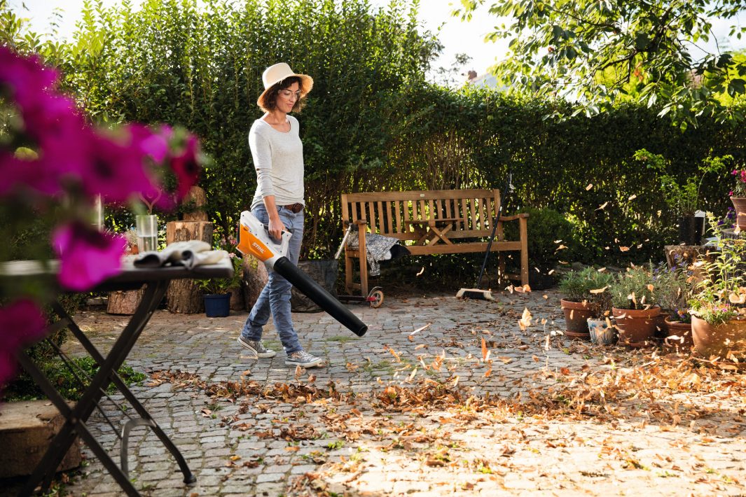 A person in a hat uses a Stihl BGA 45 blower to clear leaves from a garden patio with potted plants, bench, and greenery.