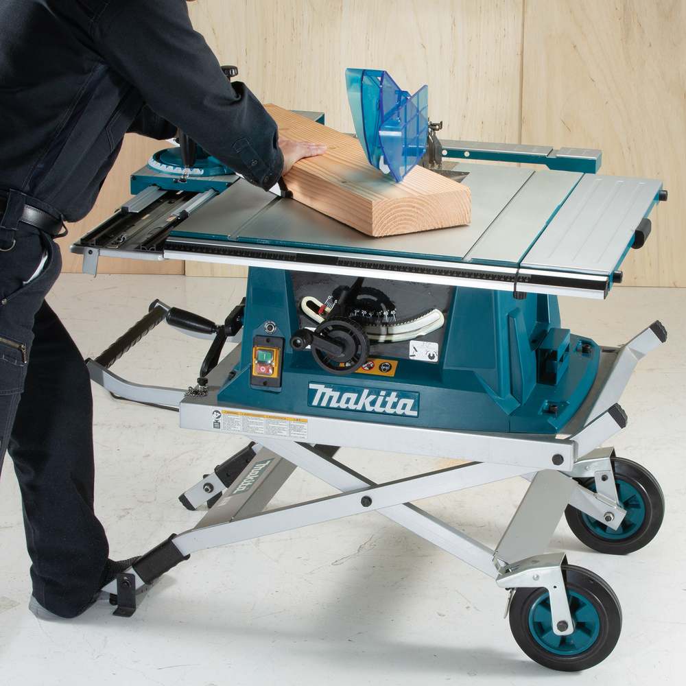 A person uses a Makita table saw and a blue push stick to cut wood; the saw sits on a wheeled metal stand against a wooden background.