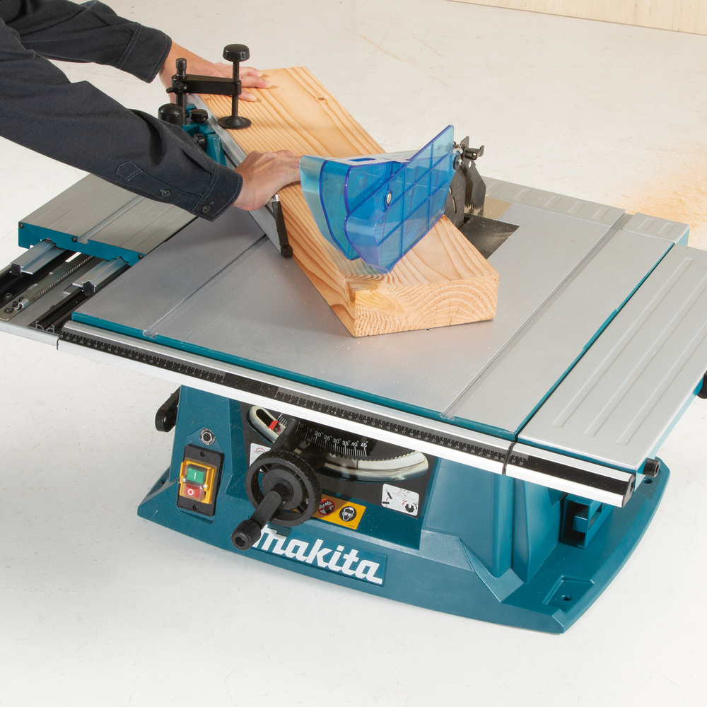 A person uses a Makita table saw to cut wood, guiding it with a blue safety guard and a push stick for protection.