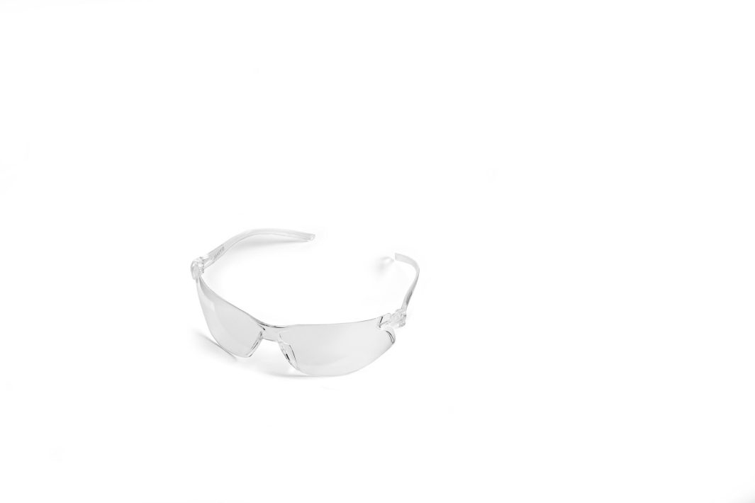 Clear safety spectacles with transparent frames and lenses rest slightly to the left on a white background in a simple, minimalist composition.
