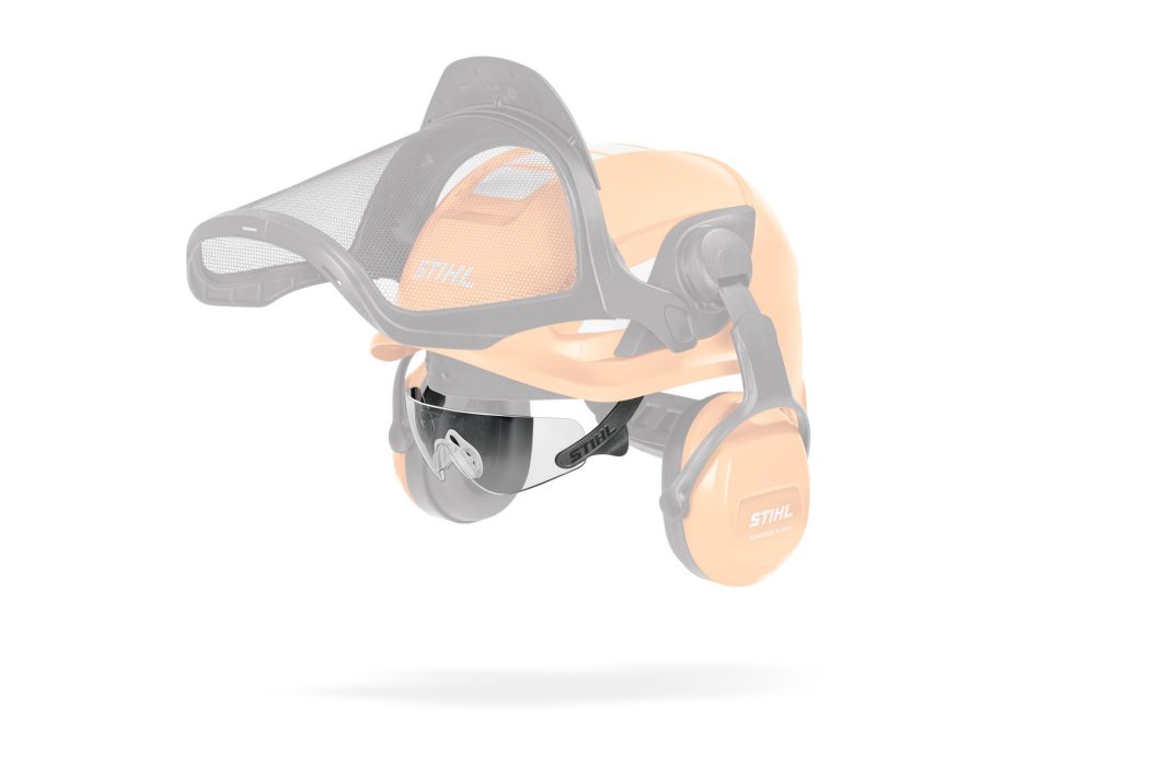 Clear face shield attachment for a STIHL safety helmet, with orange ear defenders and mesh visor partially visible in the background.