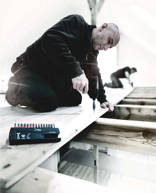 A person kneels on a wooden floor, using a power drill to fasten a board. A set of drill bits is nearby. The scene appears to be a construction or renovation project in progress.