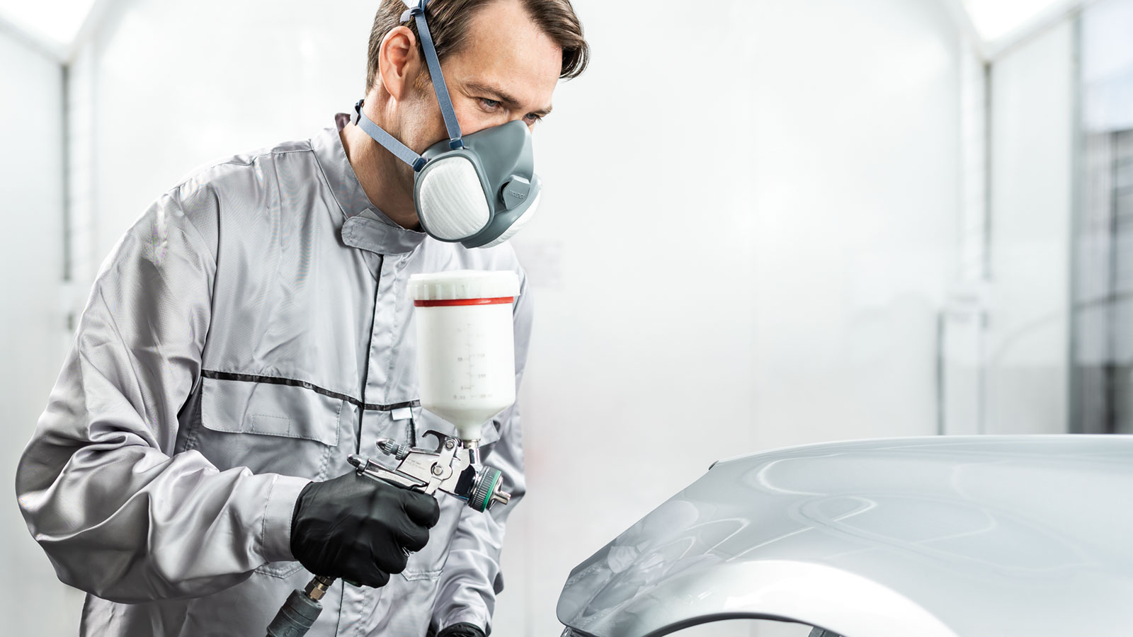 A person in protective gear and a respirator uses a spray gun to paint the bonnet of a car inside a brightly lit, professional car body shop.