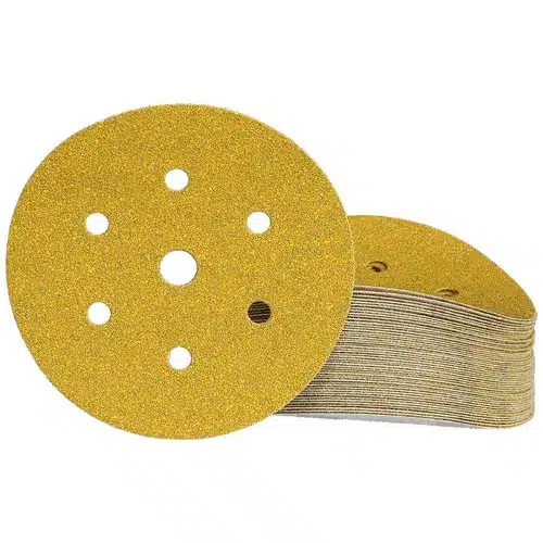 A stack of Mirka Gold 150 mm Grip 7 Holes sandpaper discs—round and gold-coloured, each featuring multiple holes—arranged with one disc standing upright in front of the rest.