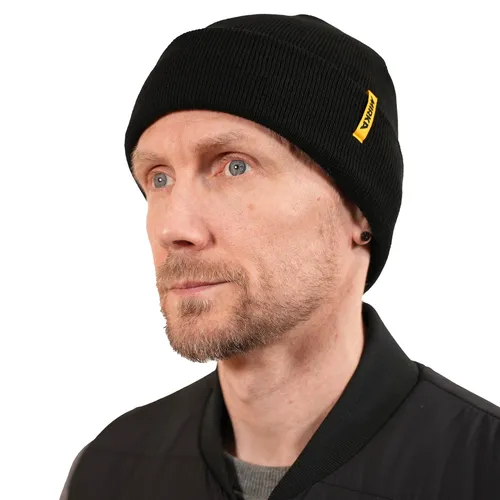 A man with a light beard and blue eyes wears a black beanie with a yellow tag and a black jacket, looking slightly to the side against a plain white background.