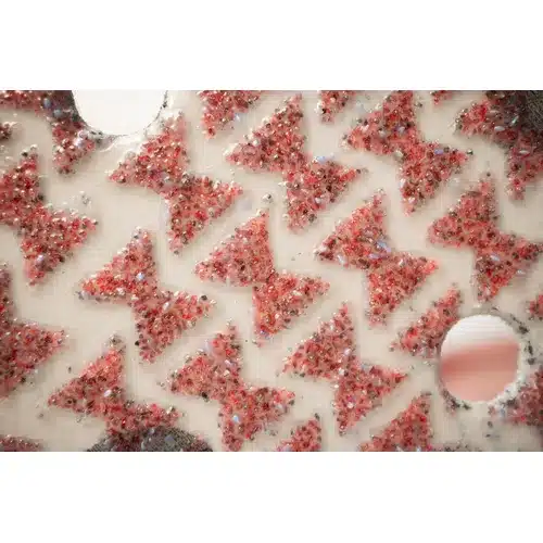 Close-up of a textured white surface featuring red, glitter-filled bowtie shapes inspired by the distinct pattern of Mirka Ultimax Ligno 150mm PSA Sanding Discs. The design is interspersed with several circular holes, echoing those found on these sanding discs.
