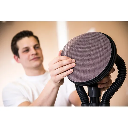 A person attaches a Mirka Abranet ACE 225mm Grip sanding disc to a round power sander, preparing the tool for use. The focus is on the hands and the sanding pad, with the person blurred in the background.