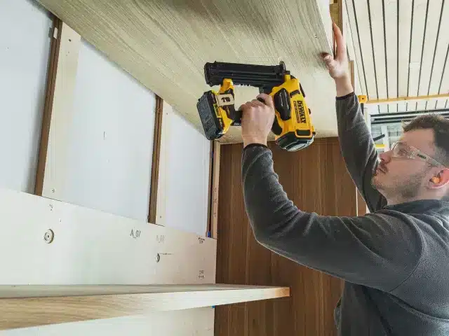 A person wearing safety spectacles uses a DeWalt DCN210SN Second Fix Cordless Nail Gun 18V Bare Unit to attach a wooden shelf to a wall cupboard.