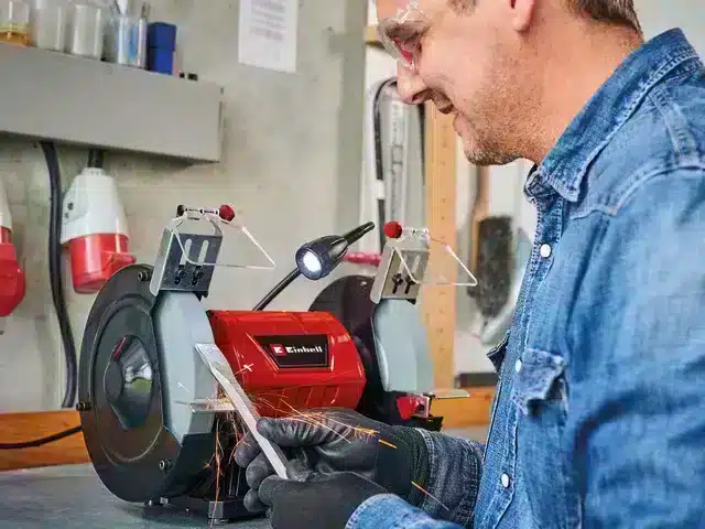 A man wearing safety spectacles and gloves uses an Einhell TC-BG 200 L Bench grinder to sharpen a metal tool, creating sparks.