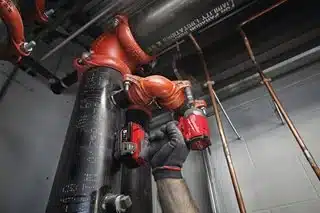 A person wearing gloves uses a Milwaukee M18FPP6L3-503B 18v 6pc Fuel Power Pack - 3 x 5.0ah Batteries cordless impact driver to secure a red metal pipe coupling onto a large black industrial pipe.