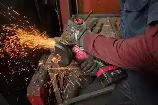 A person using a Milwaukee M18FPP6L3-503B 18v 6pc Fuel Power Pack cordless angle grinder to cut a metal bar, producing a shower of orange sparks.